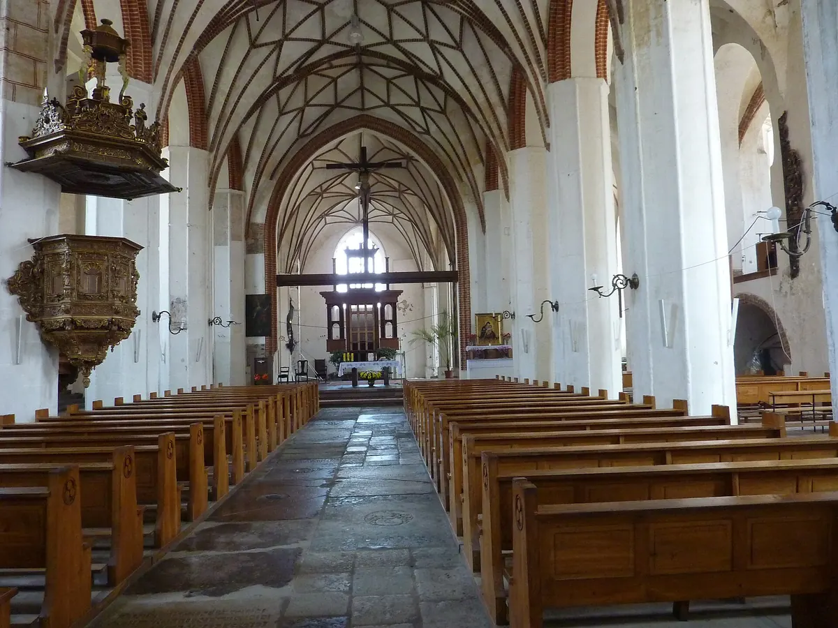 The long nave of St Catherine's Church in Gdańsk seen from the aisle: rows of wooden pews, white pillars with red brick ribs and a gilded pulpit at the left